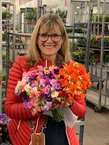 Floristin Lydia Niehüser in einem Blumenladen hält einen bunten Strauß, umgeben von Dekorationen und großen Papierblumen.