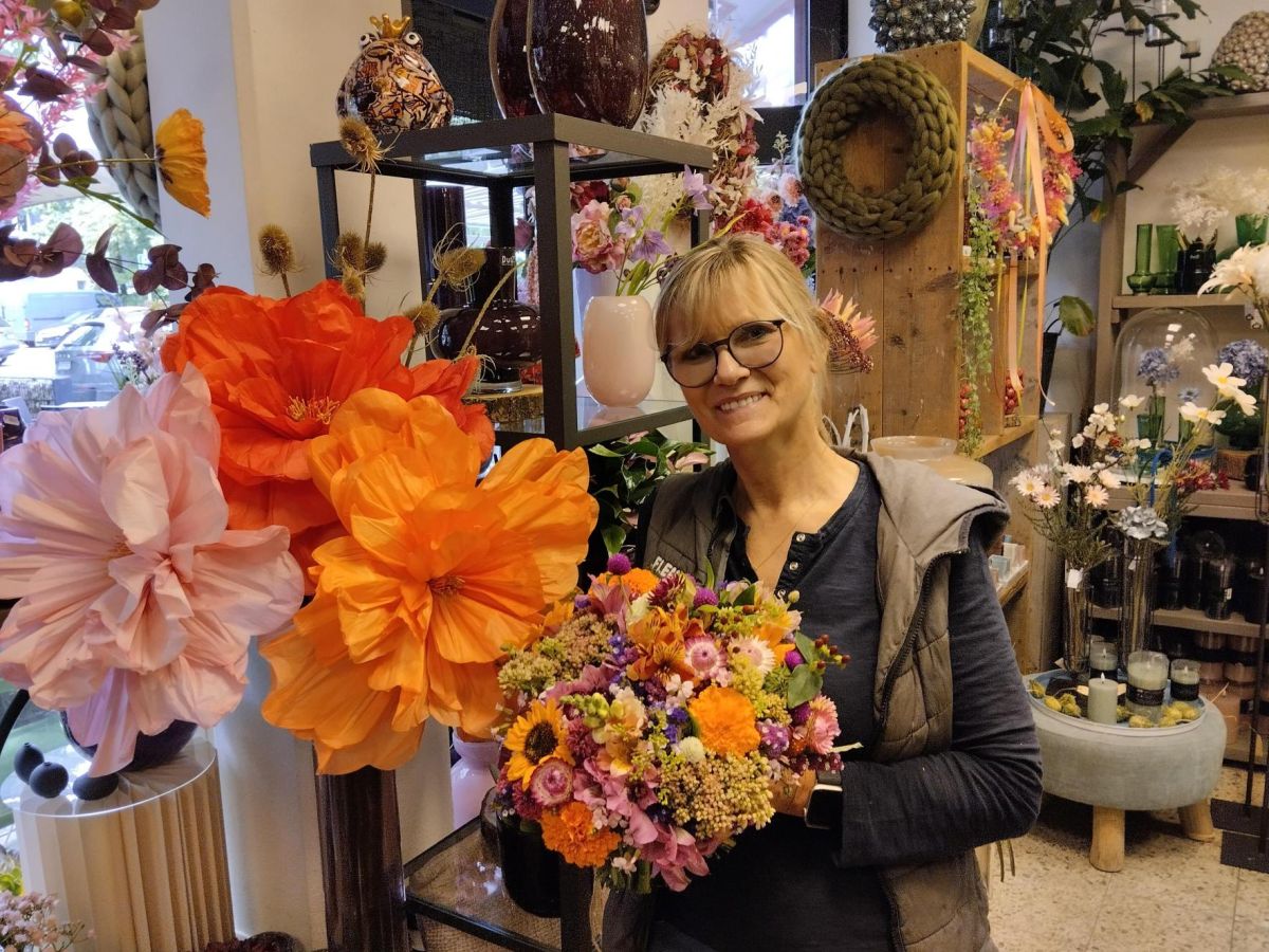 Floristin im Blumenladen hält einen bunten Blumenstrauß, im Hintergrund große dekorative Blüten und Wohnaccessoires.