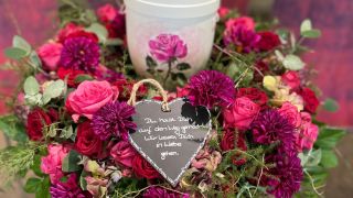 Runder Trauerkranz aus pinken und violetten Blumen mit Herzschild und Urne in der Mitte, liebevoll arrangiert.