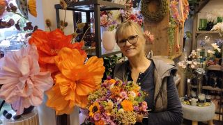 Floristin im Blumenladen hält einen bunten Blumenstrauß, im Hintergrund große dekorative Blüten und Wohnaccessoires.