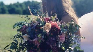 Braut im weißen Kleid hält einen üppigen Blumenstrauß in Rosa, Violett und Grün auf einer sonnigen Wiese.