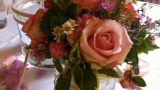 Kleines Blumenarrangement mit rosafarbenen Rosen und Grün in einem Glasgefäß auf festlich gedecktem Tisch.